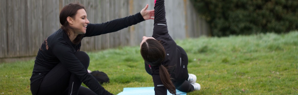Gemma kneeling on the grass beside a client on a mat, smiling with one arm raised as she coaches the stretch.